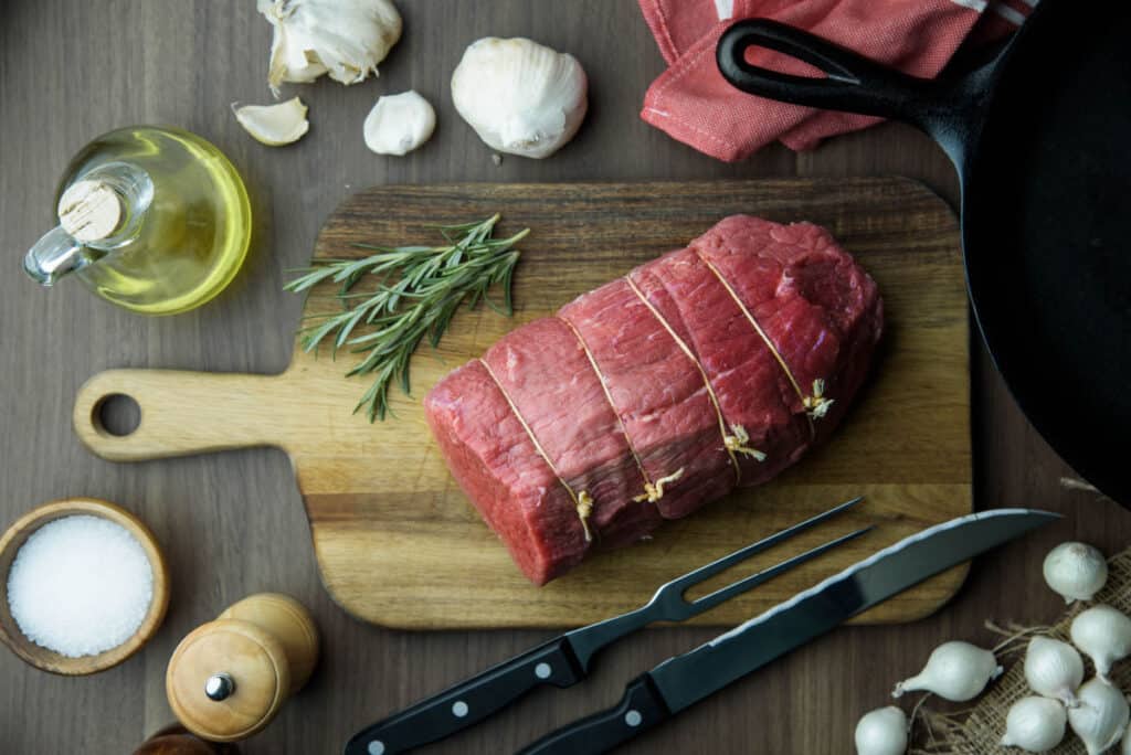 A raw eye of round roast, tied and sitting on a paddle shaped board, surrounded with garlic and he.