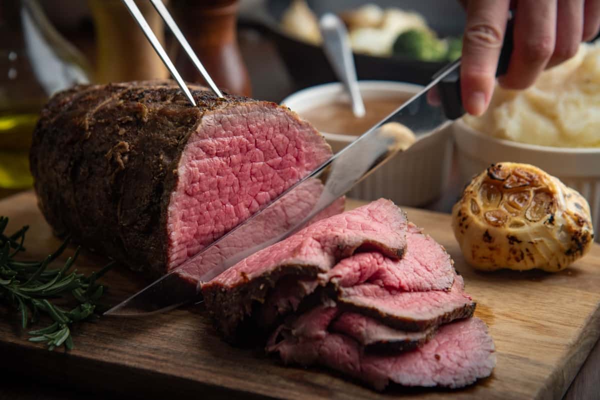 slicing roasted eye of round beef with knife, on cutting board with roasted garlic and a rosemary sprig