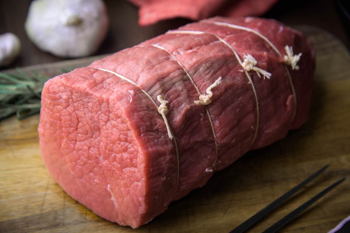 raw eye of round roast tied with butchers twine, on a cutting board with soft focus garlic and rosemary in the backgro.