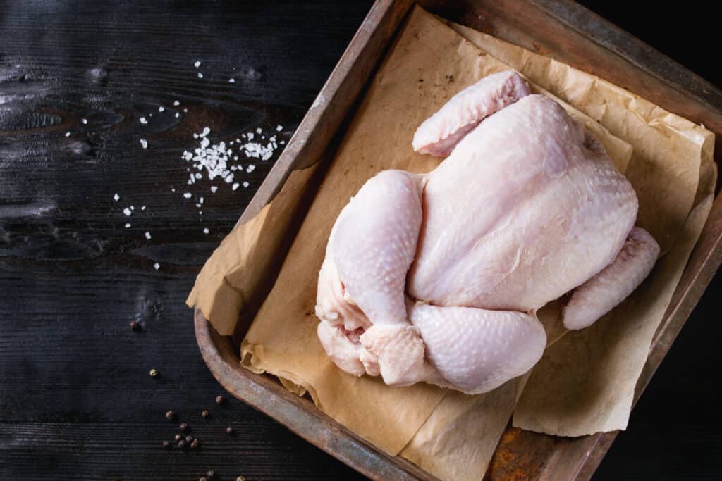 A whole chicken on parchment paper in a baking tray, next to some loose salt on a table