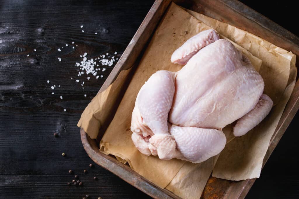 A whole chicken on parchment paper in a baking tray, next to some loose salt on a ta.