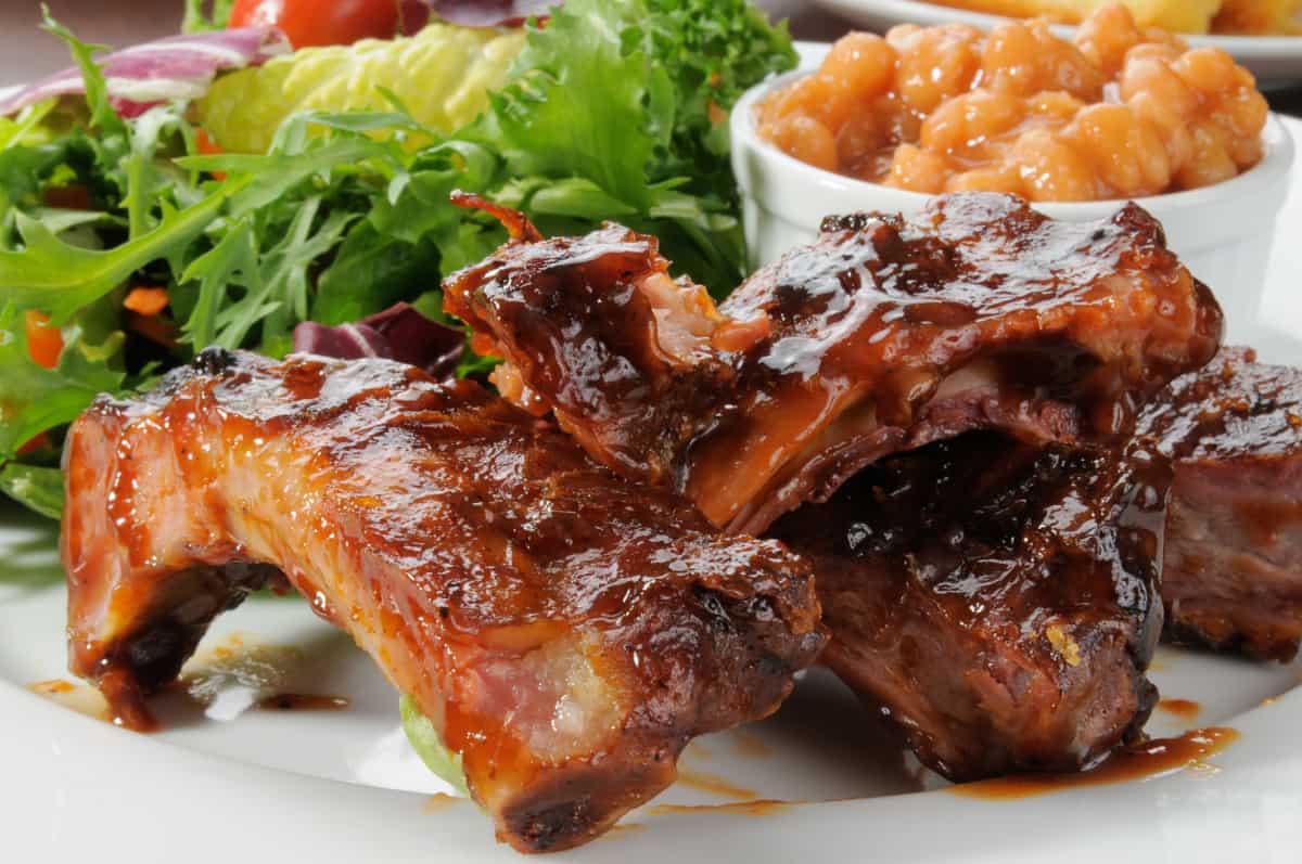Close up of baby back ribs, slathered in sauce, in front of a pot of beans and some green salad