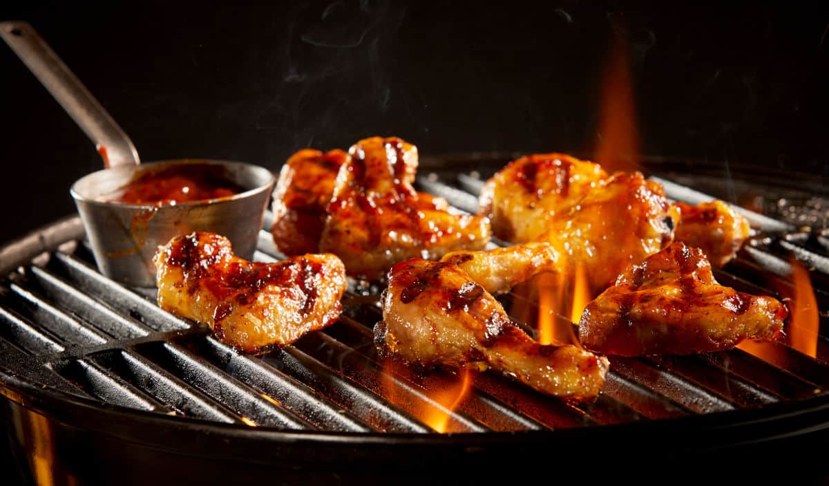 Marinated chicken wings, and a pot of sauce, being warmed on a BBQ gr.