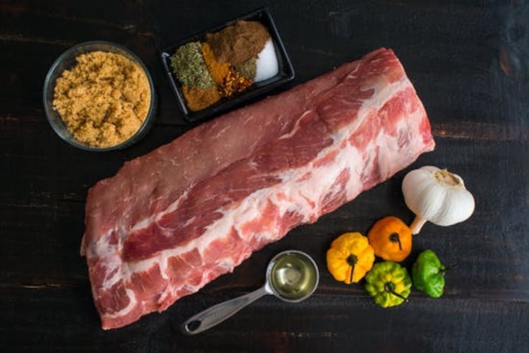 Raw Crowd Cow baby back ribs, on a dark table with herbs, spices, some chili peppers and a bulb of garlic