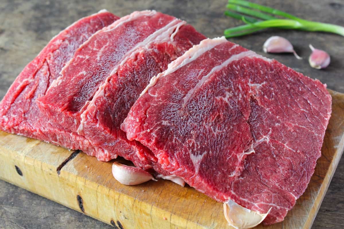 Four slices of raw top round steak on a wooden cutting board, surrounded by a handful of garlic clo.