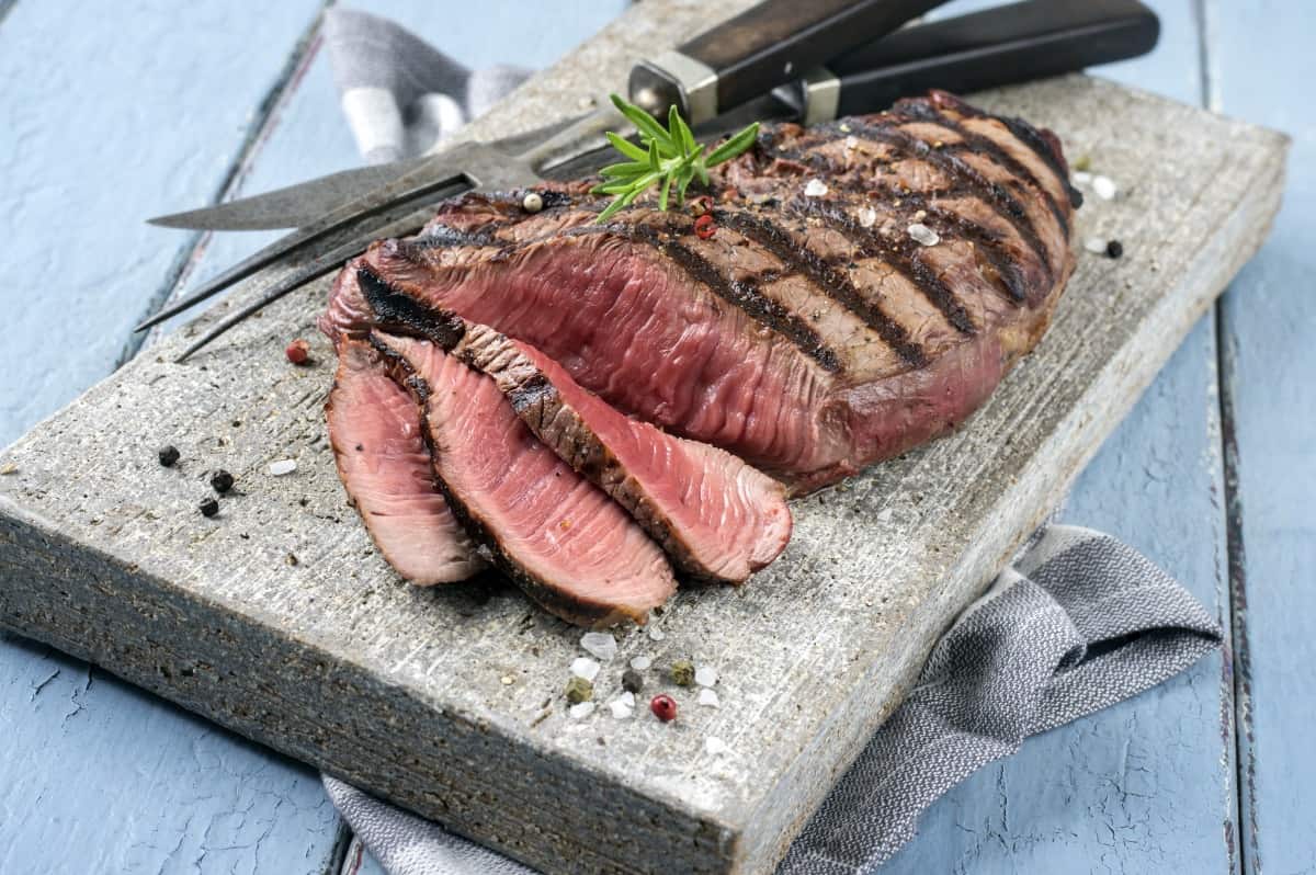 sliced grilled top round steak on a stone platter, with a carving knife and fork and a sprig of rosemary on .