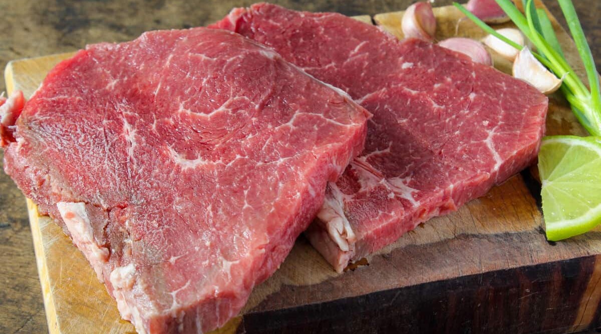 Two, raw top round steaks on a cutting board with garlic, lime and spring onion.