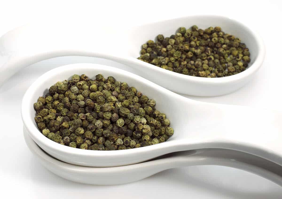 Two white ceramic spoons full of green peppercorns on a white surface