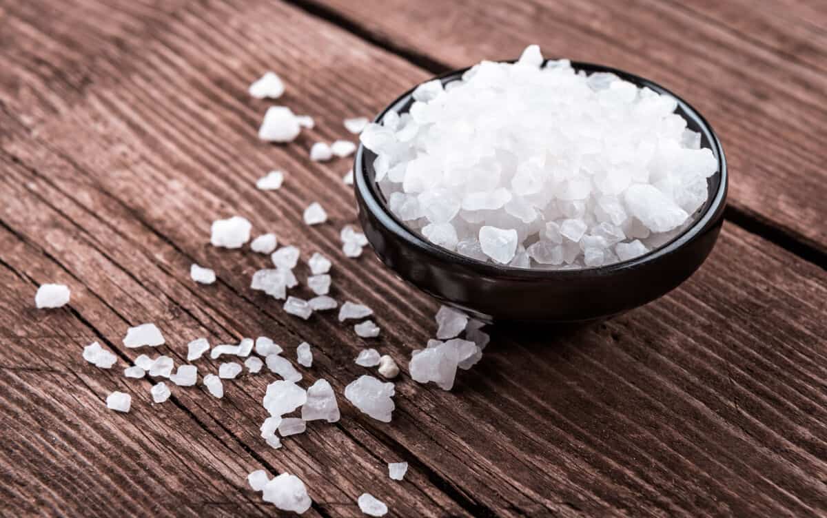 A bowl of sea salt with a few flakes surrounding it on a table