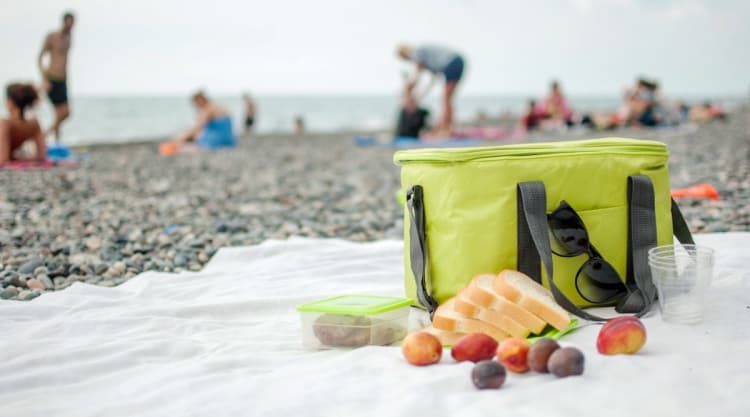 A soft-sided cooler bag, on a towel on a beach, with bread and fruit beside it.