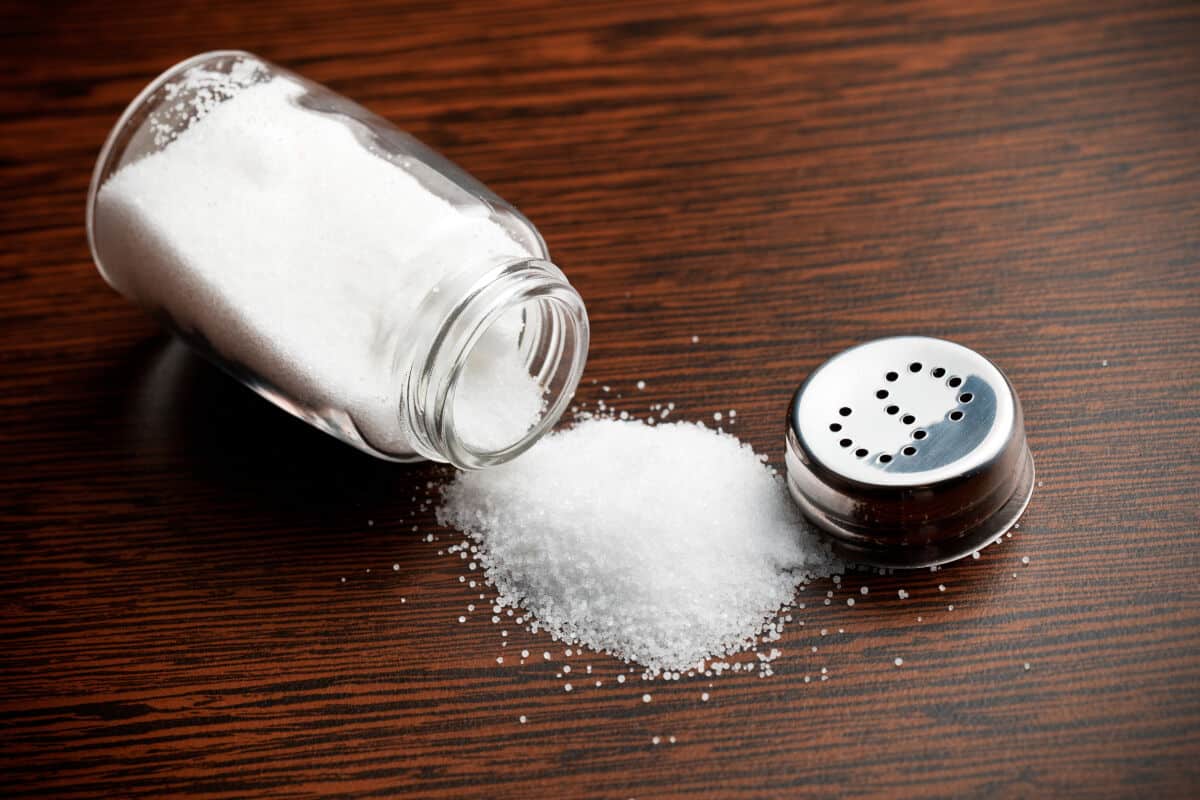 A shaker of table salt on it's side, lid off, some spilled onto the ta.