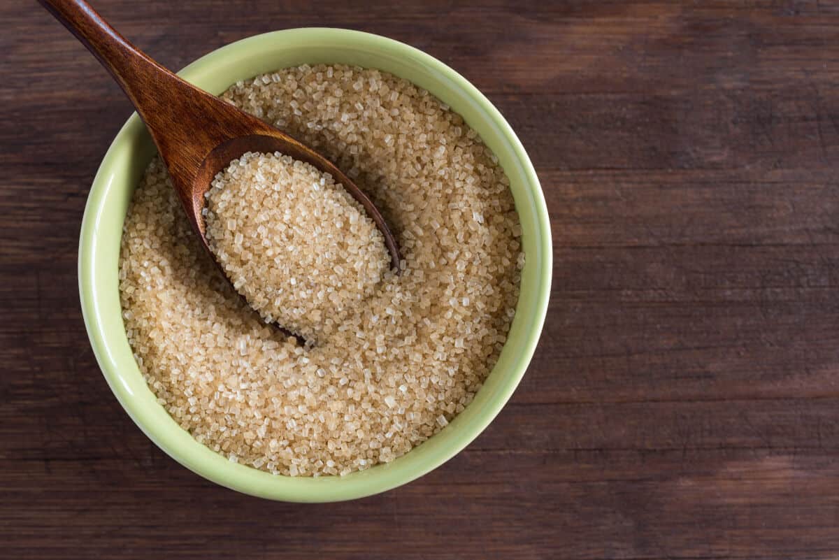 A green bowl full of turbinado sugar, with a wooden spoon in, sitting on a dark wooden ta.