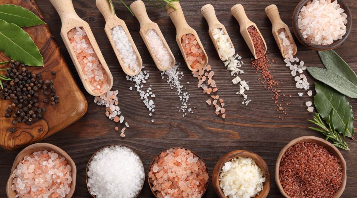Different types of salt in bowls, in scoops, and spread out on a wooden table.