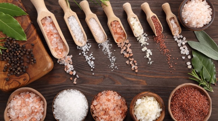 Different types of salt in bowls, in scoops, and spread out on a wooden table