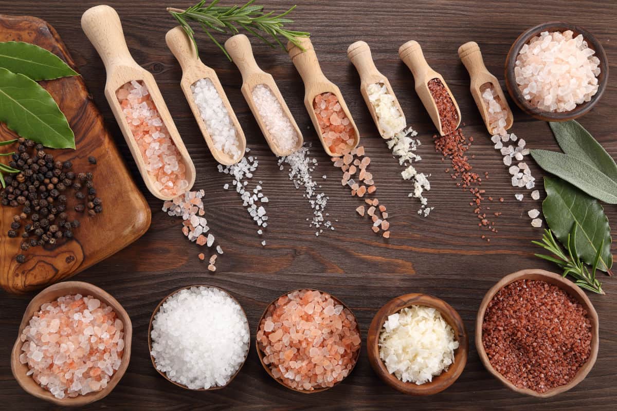  Different types of salt in bowls, in scoops, and spread out on a wooden ta.