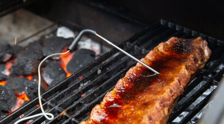 Ribs on a charcoal grill, with a thermometer probe attached.