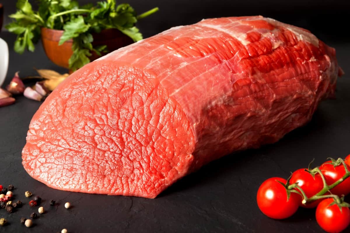 Raw eye of round roast on dark surface with tomatoes and some gre.