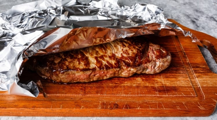 A steak resting under foil on a cutting board.
