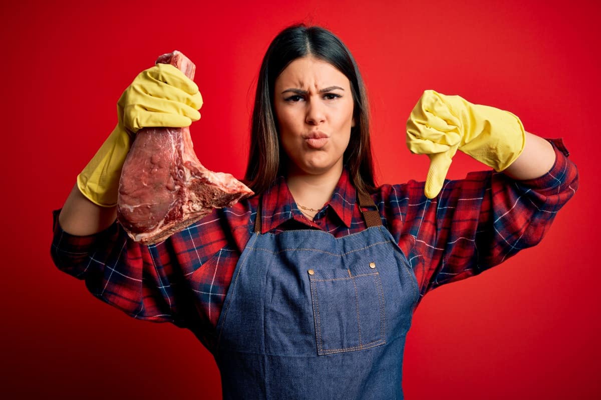 A woman in yellow gloves holding a gone bad steak, making a thumbs down gest.