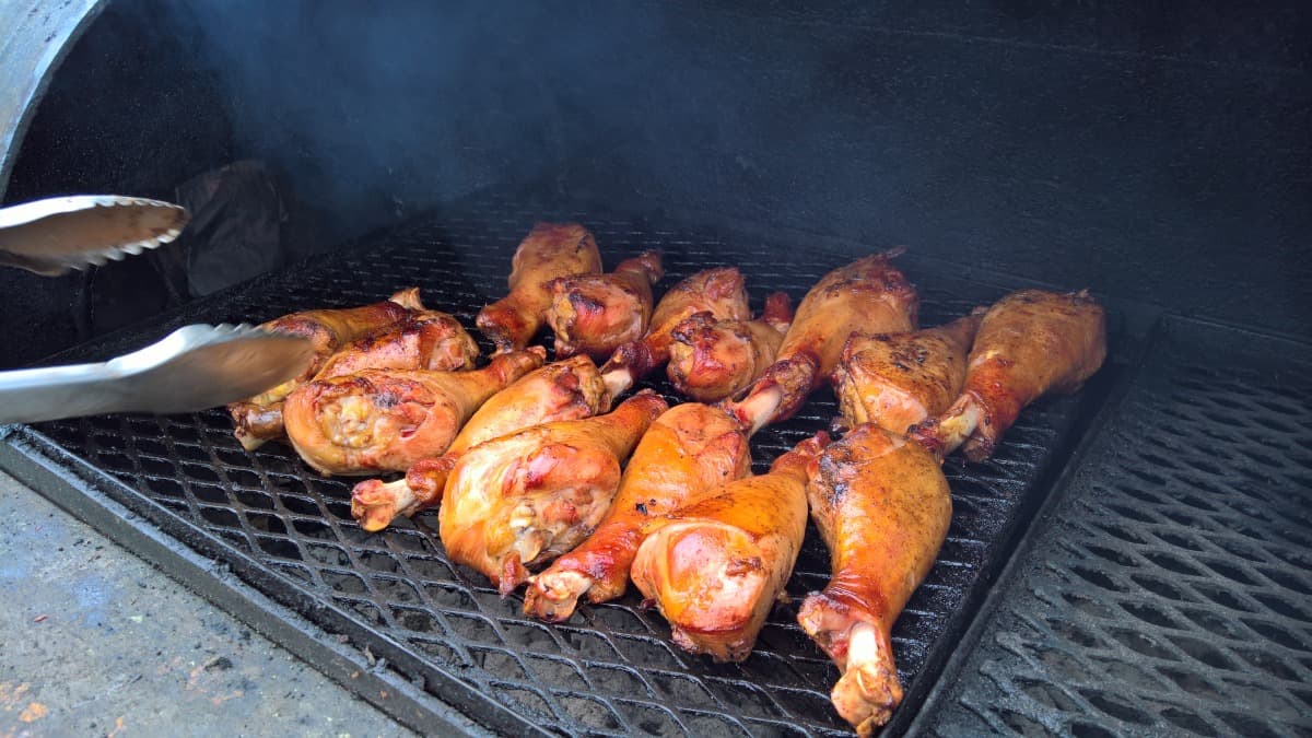 Turkey legs in a smoker being turned by tongs in a black gloved h.