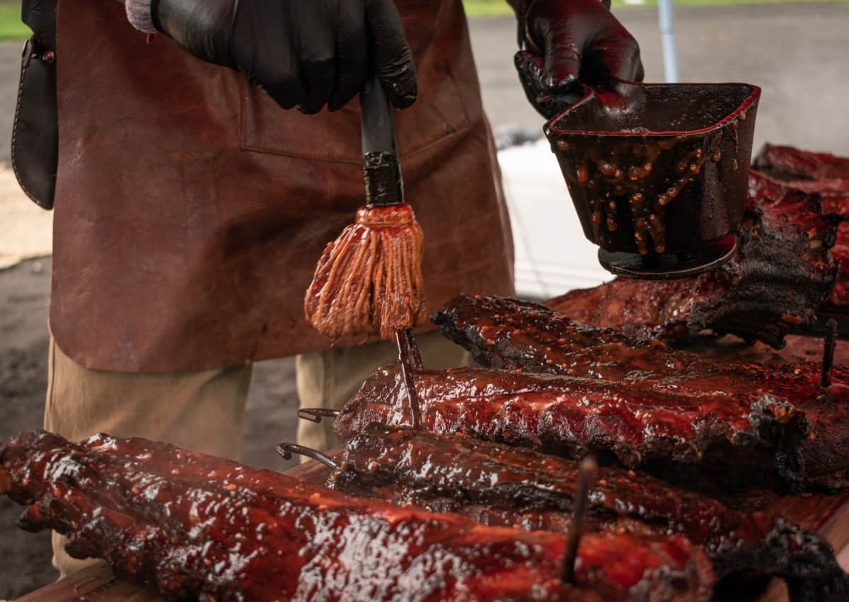 Many racks of ribs being mopped with sauce, by a man in a leather ap.