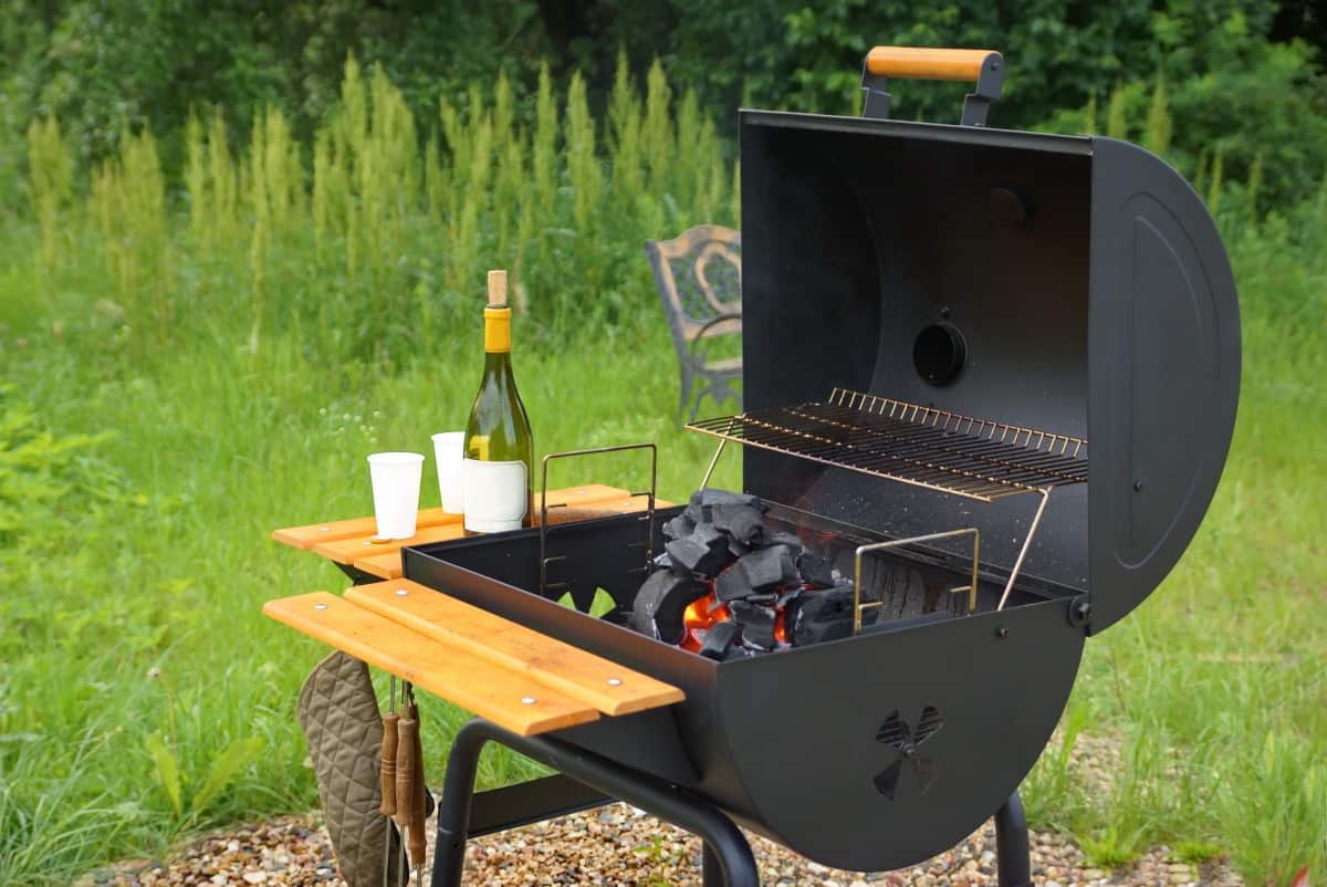 A barrel shaped charcoal grill with wooden side and front shelf, lid open and champagne on one side.