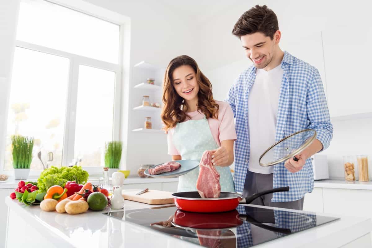 Man and woman dropping a large steak into a frying pan, in a well lit kitchen with fruit and veg laying aro.