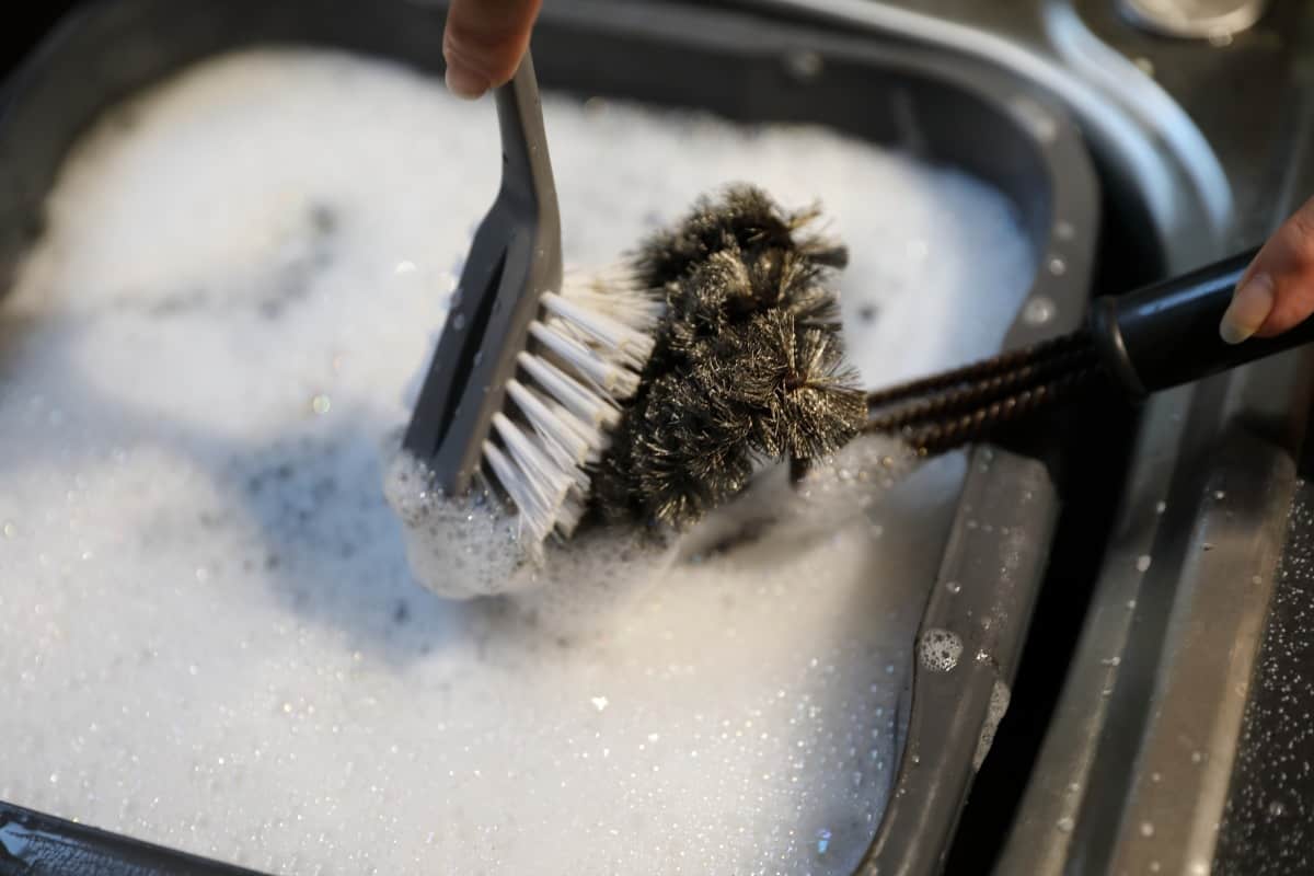 A metal grill brush being cleaned with a nylon dish brush in a bowl of soapy wa.