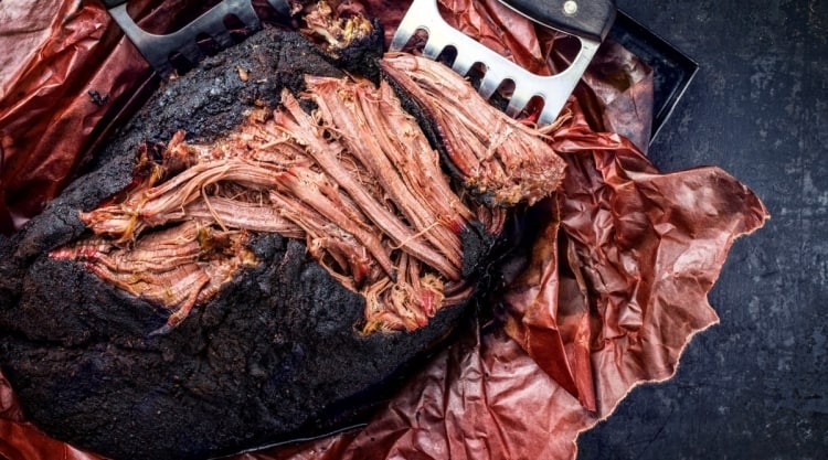Smoked pork butt on butcher paper with two meat claws, the top part having been pulled and shredded.