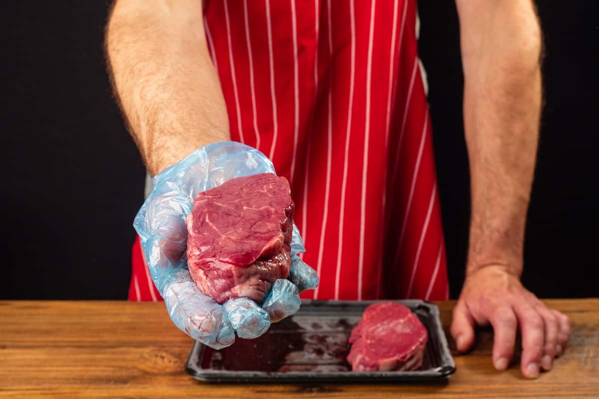 A man in red apron holding up a raw filet steak to the cam.