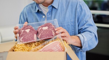A man unpacking a mail order steaks delivery, showing two ribeyes, some bacon, and a box with straw like packaging