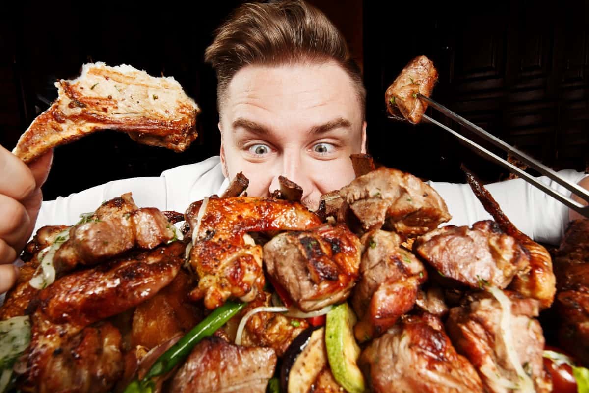 A man looking up over a large pile of grilled BBQ meat, so you can only see his nose up, with a piece of meat in each hand.