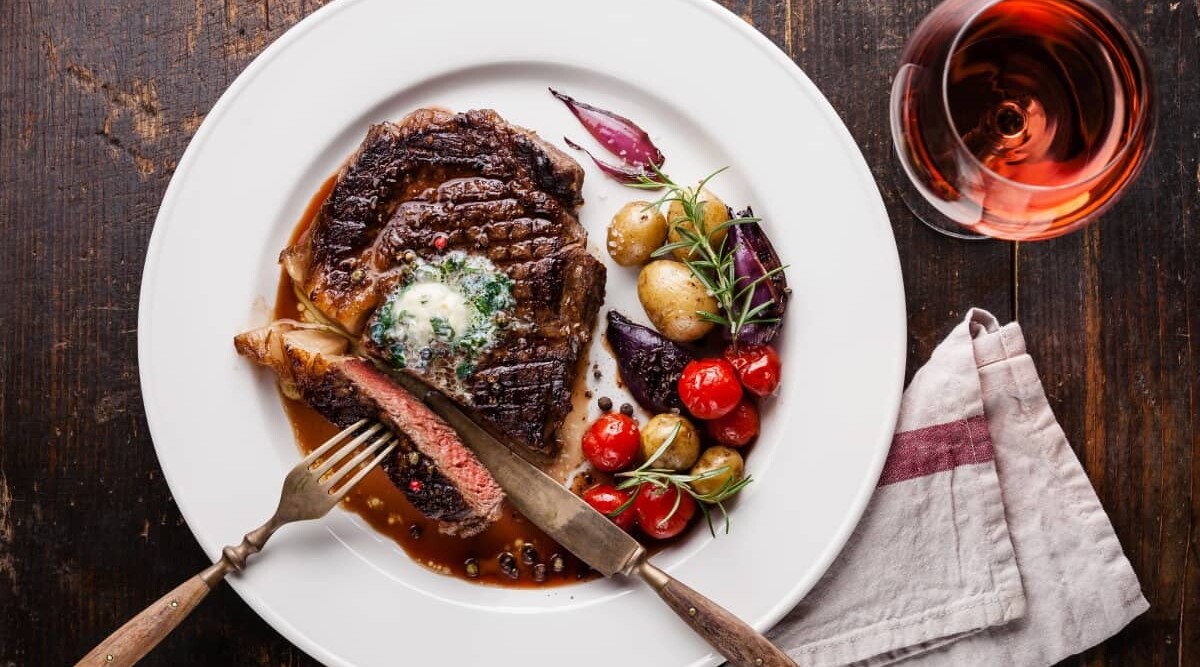 An overhead shot of reheated steak on a white plate, with some roasted veg, next to a glass of wine.