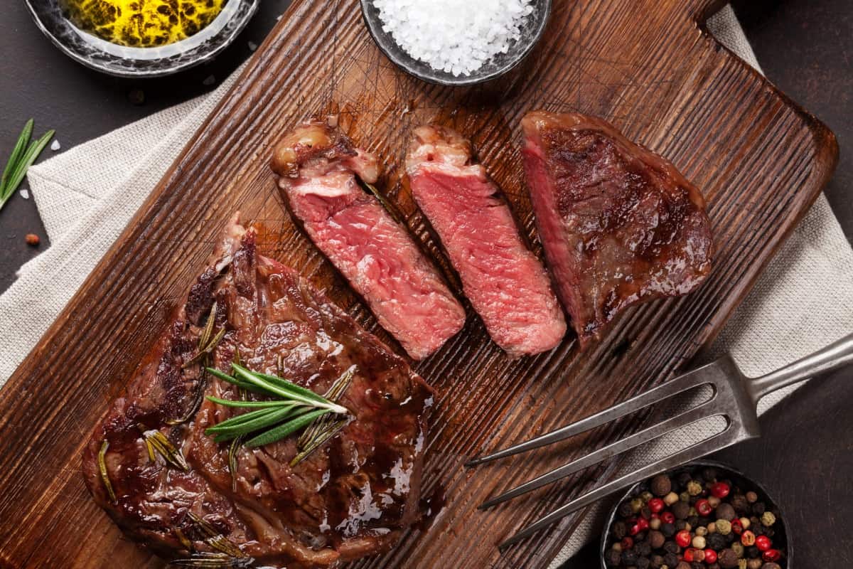 sliced NY strip on a wooden cutting board, with rosemary sprig and two pots full of rock salt and pepperco.