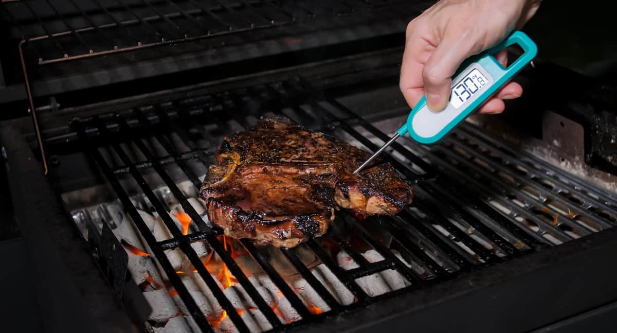 A steak on a charcoal grill, with a blue and gray instant read thermometer inserted.