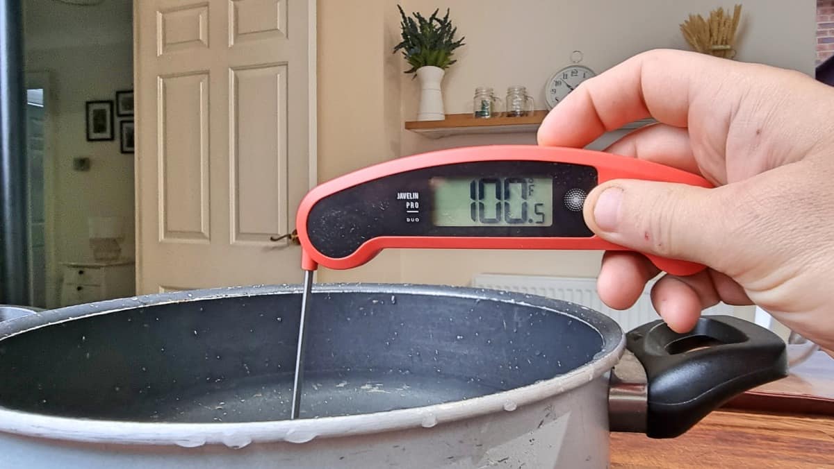 An instant read thermometer showing a temperature of 100 F with the tip in a pan of water