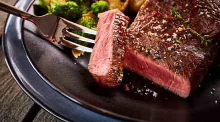 A thick steak with a slice cut away and being held on a fork, in front of some veg on a plate.