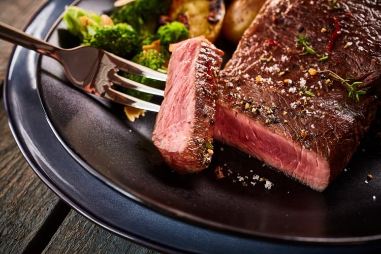 A thick steak with a slice cut away and being held on a fork, in front of some veg on a plate.