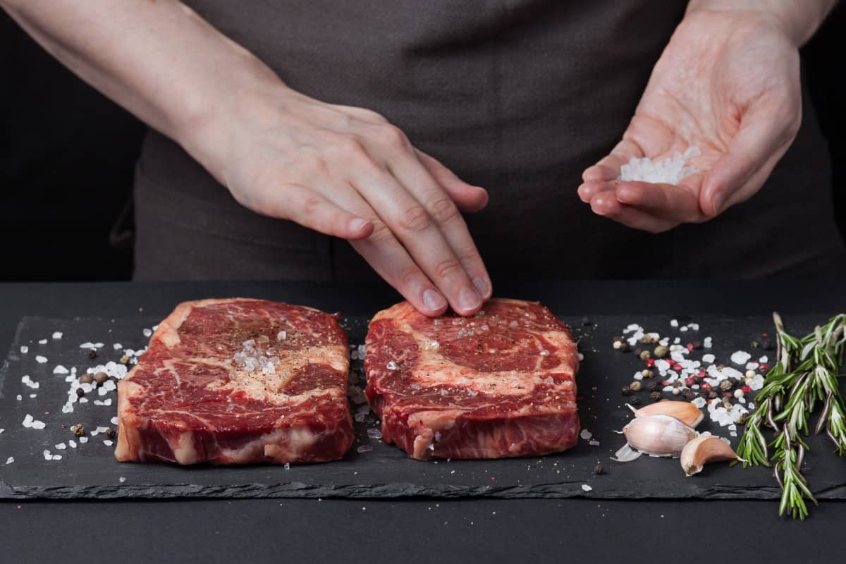 A female chef salting two steaks on a slate board, next to garlic and rosemary sprigs