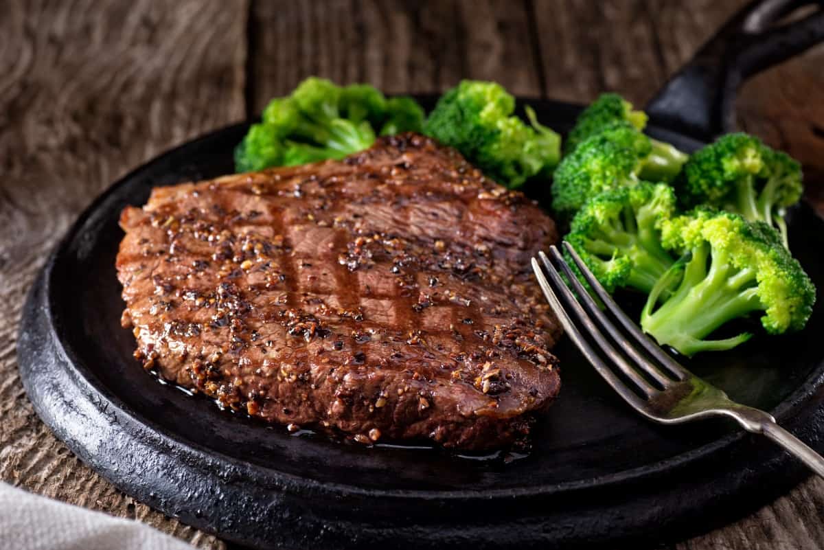 A grilled peppered steak, on a black plate next to some boiled broccoli