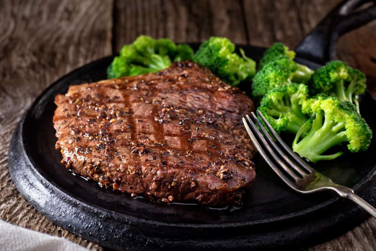 A grilled peppered steak, on a black plate next to some boiled brocc.