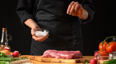 A man in black apron, sprinkling salt over a steak on a cutting board.