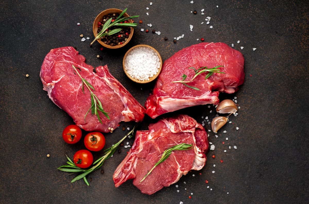 Three raw steaks on a cutting board with salt, herbs, and spi.