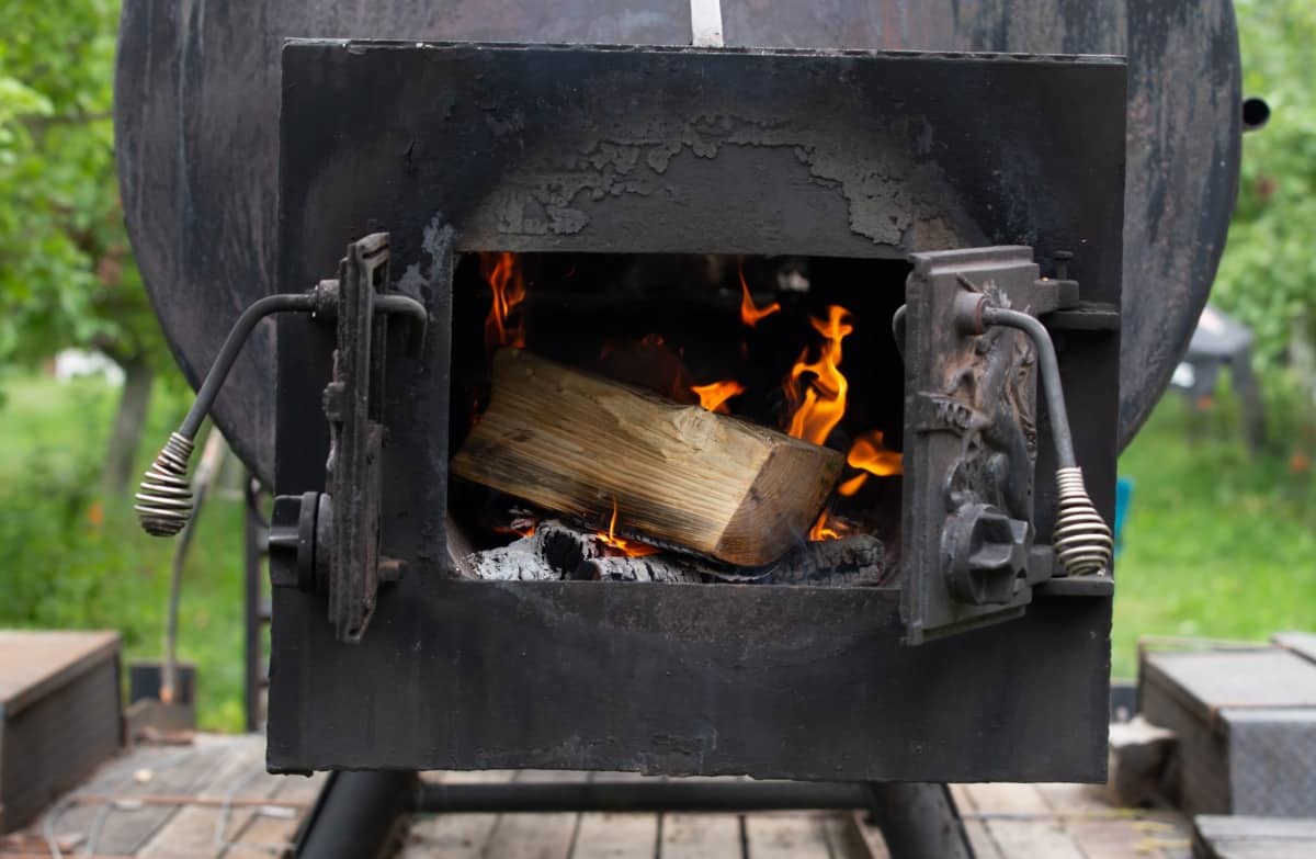 A log of wood seen burning through the door of a firebox in an offset smoker