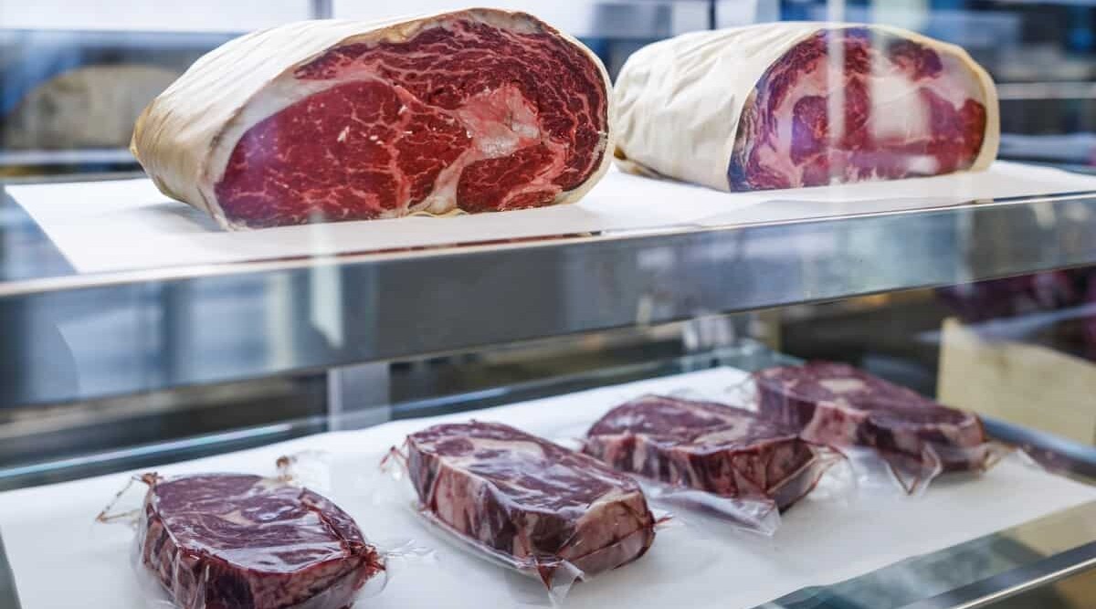 Some cuts steaks, and some whole joints, wrapped in clear plastic in a glass refrigerator.