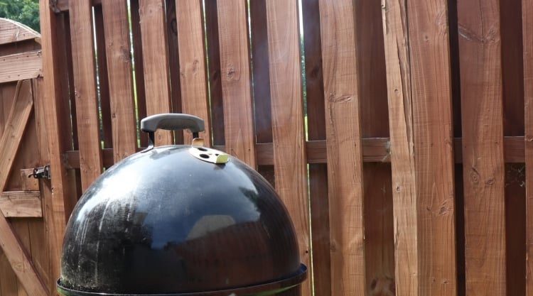 Thin blue smoke coming out of the top vent of a Weber Smokey Mountain cooker, in front of a wooden fence