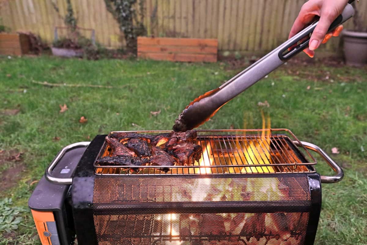 A womans hand turning lamb cutlets with tongs on a Biolite firepit+.