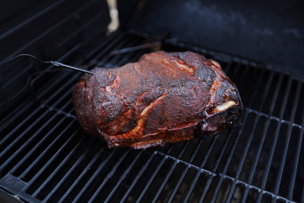 Smoked pork shoulder on cast iron grates, with a temperature probe inserted and a trailing wi.