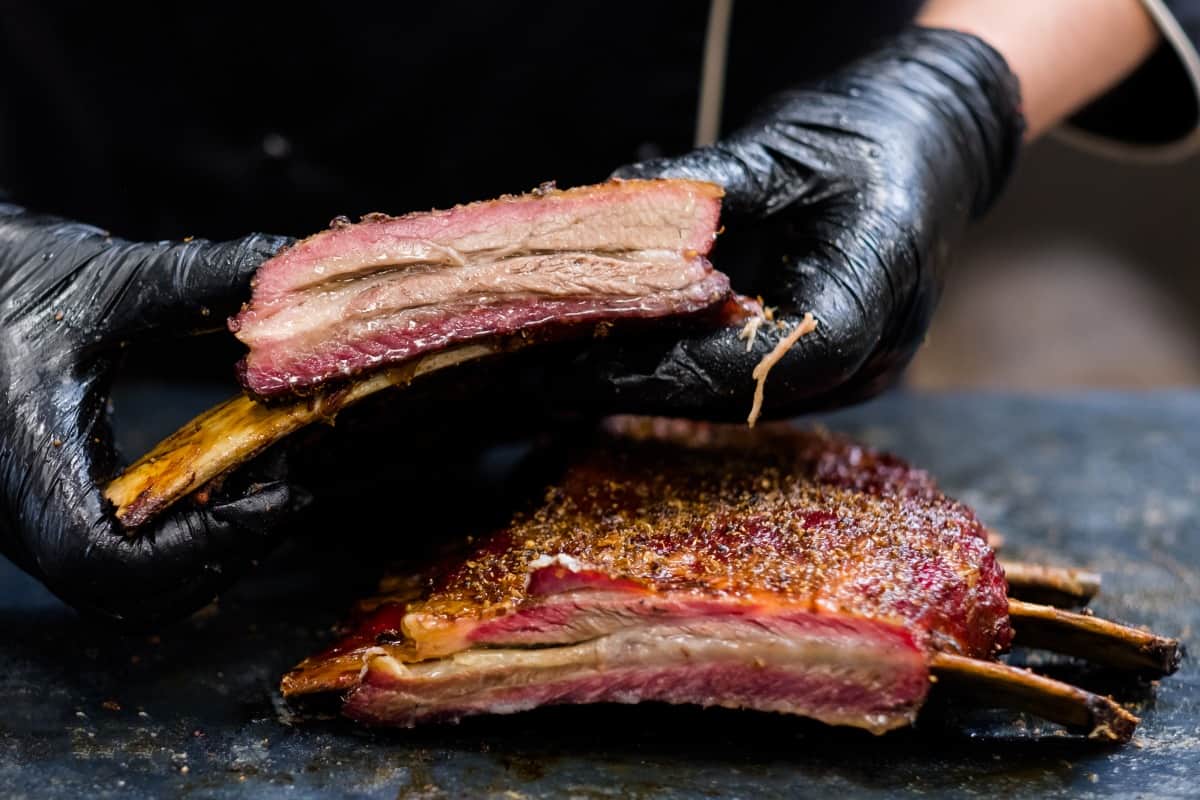 Smoked ribs, sliced, with one held up by a mans hands in black gloves.