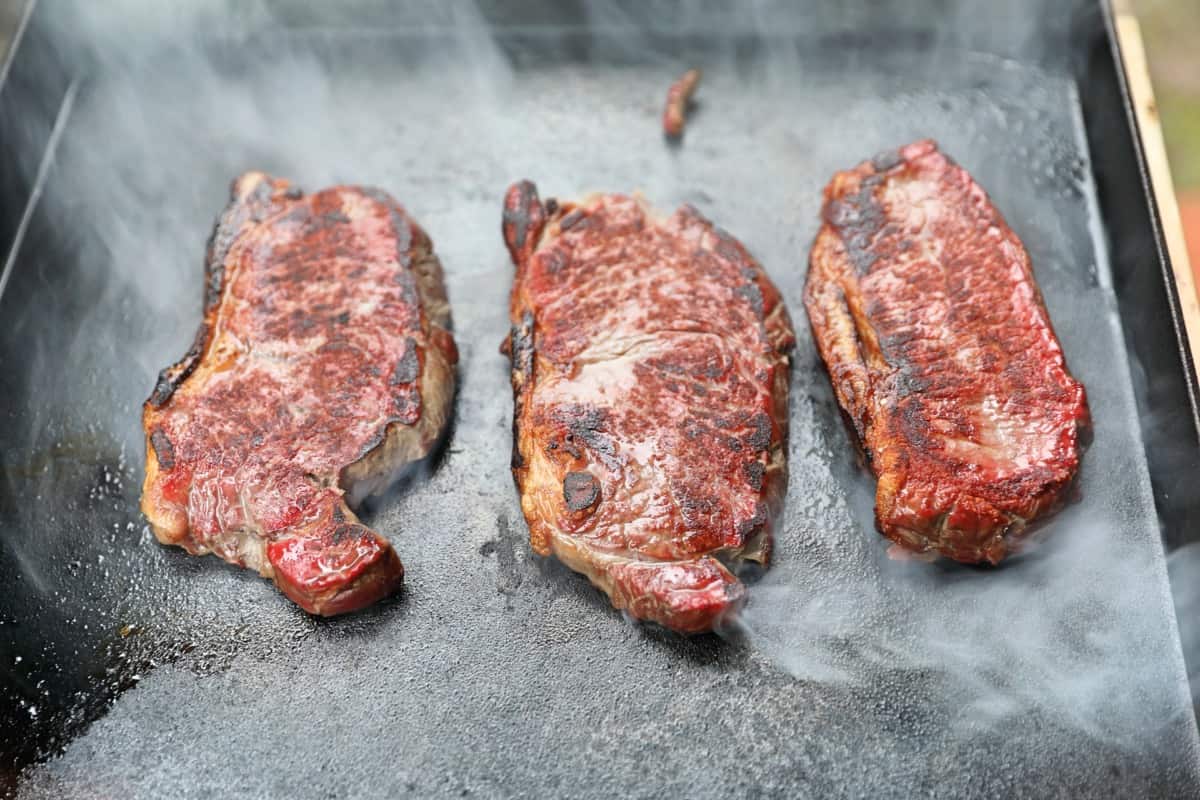 3 steaks searing on a cast iron griddle.